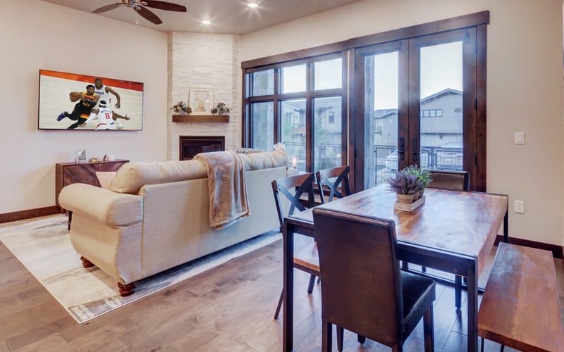 Open-plan living and dining room with beige sofa, fireplace, dining table, and floor-to-ceiling windows overlooking city buildings.