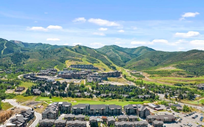 Aerial view of mountain resort town with residential buildings, forested slopes, and green valleys under blue sky