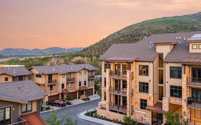 Mountain resort community with multi-story stone and stucco homes at sunset, forested hillside and valley beyond.
