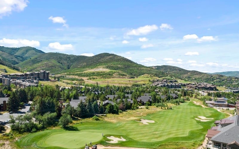 Golf course with manicured fairways and sand bunkers, surrounded by forested residential area and green mountains under blue sky