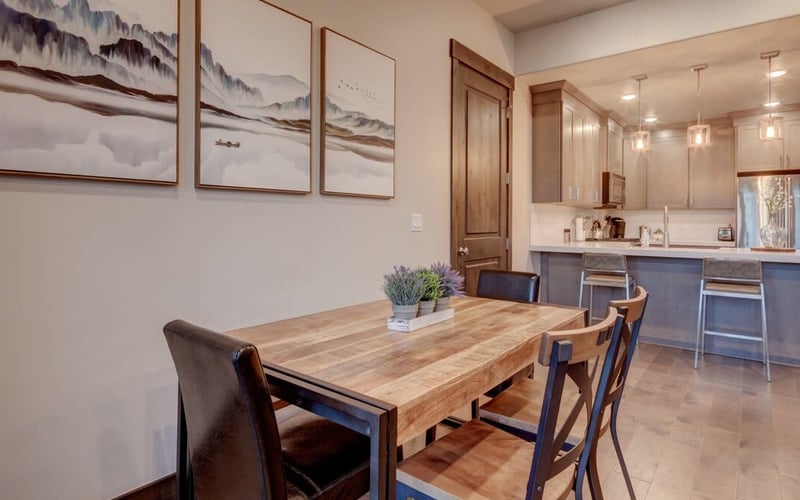 Open-plan dining and kitchen area with wooden table, upholstered chairs, and framed mountain landscape artwork on walls