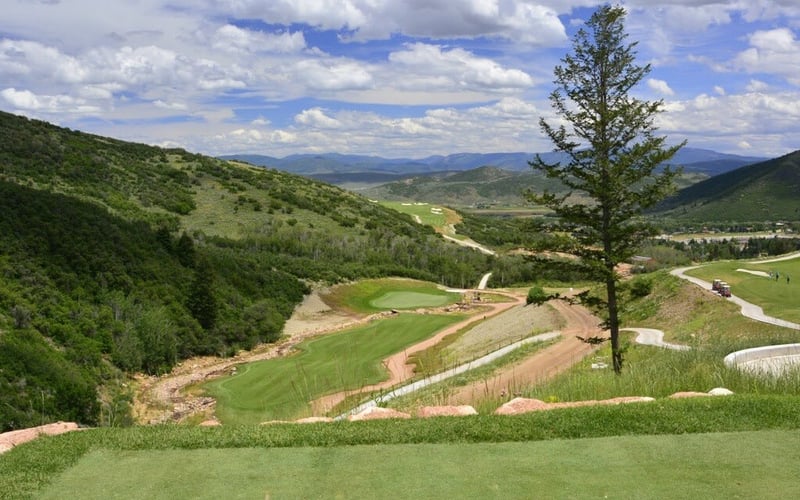Hillside golf course with manicured greens, forested slopes, and distant mountains under partly cloudy sky