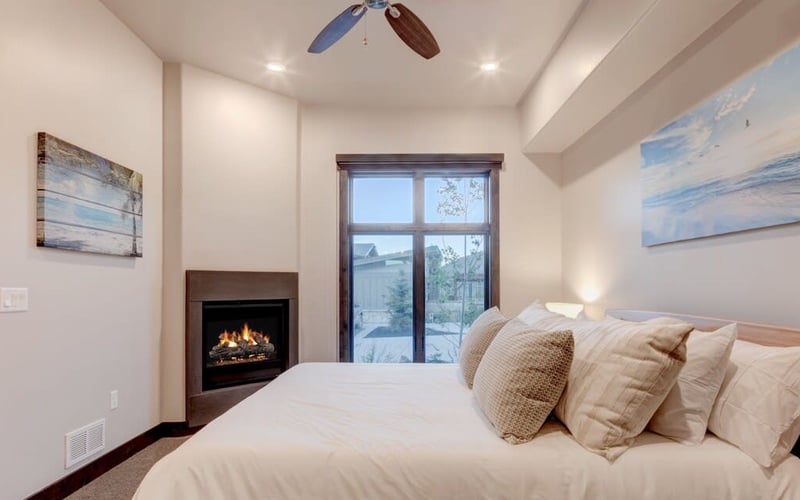 Master bedroom with fireplace, ceiling fan, large bed with decorative pillows, and glass doors opening to a deck with mountain views