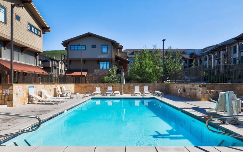 Resort swimming pool with lounge chairs, stone patio, and multi-story residential buildings under clear blue sky