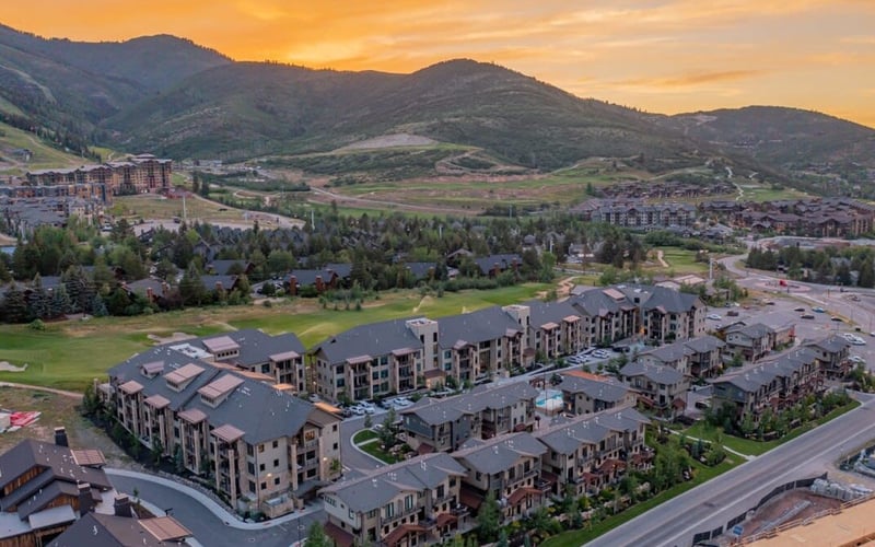 Aerial view of residential complex with multi-story apartments in mountain valley at sunset, surrounded by forested hills and green fields.