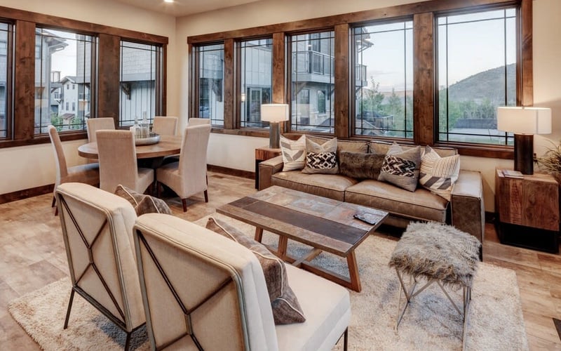 Open-plan living and dining room with wood-framed windows, mountain views, beige seating, and wood coffee table.